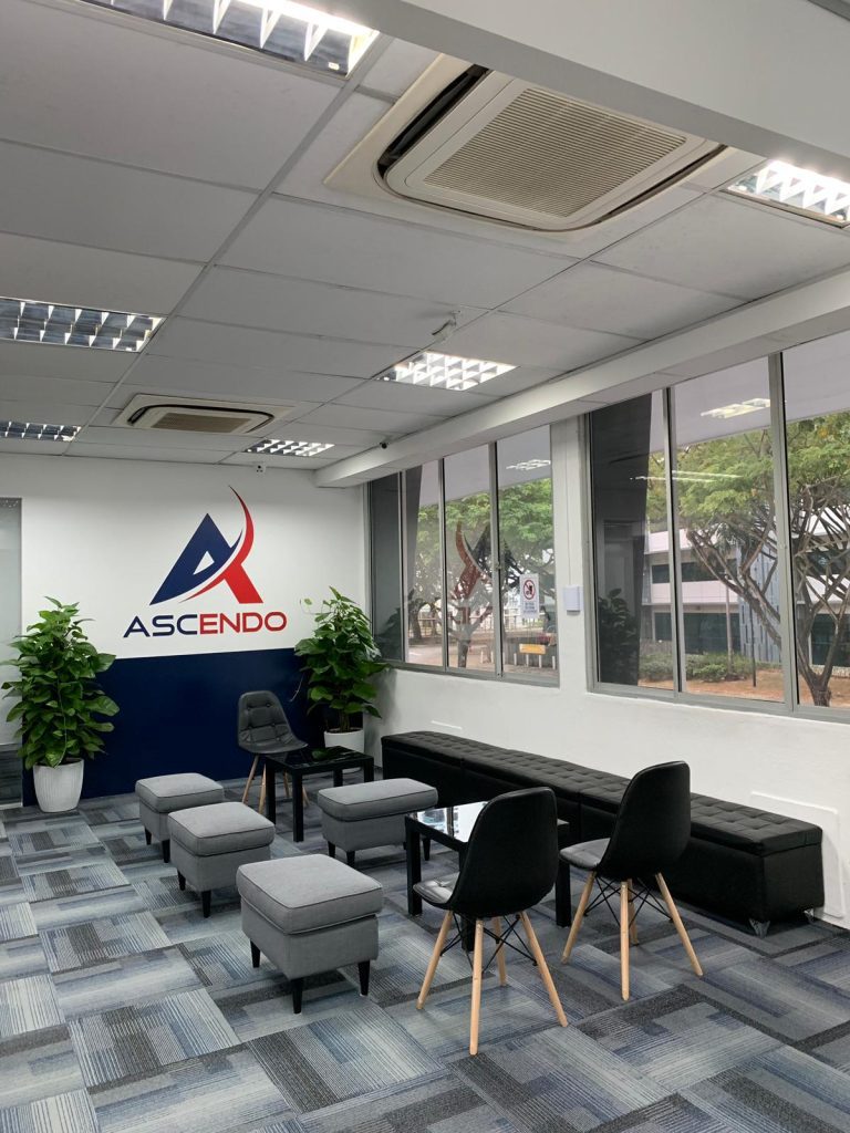 A reception area with the Ascendo logo on a navy feature wall, grey carpet, low grey stools and black chairs, and windows along one side.